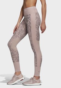 Žena-modelka nosí vysoké legíny v barvě švestkové s hadím vzorem a sladěné švestkové sportovní boty s bílými podrážkami.