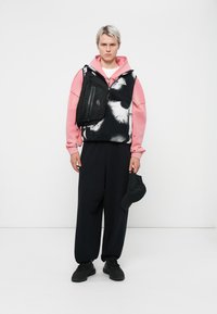 Modelis ģērbies rozā hoodie, melnā un baltā rakstainā vestē, brīvās melnās biksēs un melnās kurpēs, tur rokās melnu cepuri un mazu soma.
