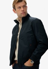 Chaqueta bomber azul marino con cuello alto, cierre frontal y bolsillos laterales. Tela suave con puños y dobladillo de canalé. Llevada sobre una blusa de un tono crema claro.