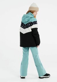 Veste de ski colorée avec une base noire, capuche bleu canard, motif en chevron blanc et marine, accents floraux, associée à un pantalon de ski bleu clair et des bottes noires.
