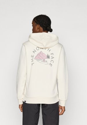 Sudadera con capucha de color crema con un gráfico de montañas y árboles en la parte trasera, con el nombre de la marca "THE NORTH FACE" curvado alrededor del diseño.