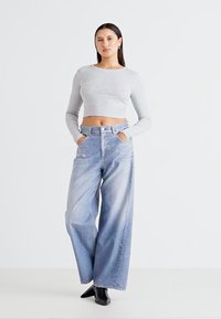 Siv prugasti dolgi rokav skrajšani top, skladen z svetlo modrimi širokimi jeansi. Model stoji z rokama v žepih v minimalističnem okolju.