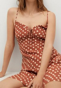 Ruskea pilkullinen mekko, jossa on sä ädettävät spagettiolkaimet, röyhelöreunat, keskellä solmimistiedot ja vartalonmyötäinen muoto. Sujuva kangas.