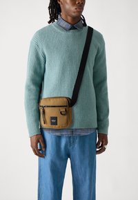 Tan crossover taske med lynlåslukning, en frontlomme og et logo-patch, parret med en tekstureret lyseblå sweater og blå denimbukser.