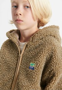 Bambino biondo che indossa una giacca in pile con zip di colore beige, con cappuccio e una piccola toppa colorata sul petto.