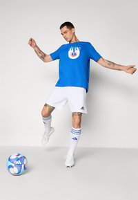 T-shirt en coton bleu avec un graphisme "ITALIE", shorts blancs, chaussettes blanches à rayures bleues, et un ballon de football bleu et blanc à motifs sur le sol.