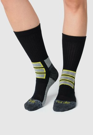 Chaussettes noires et grises avec des accents rayés jaunes vif. Texture côtelée avec des blocs de couleur contrastants sur les côtés et des zones de renforcement au niveau des orteils.