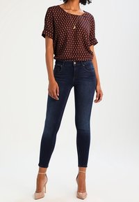 Kvinna iklädd en brun mönstrad kortärmad blus med mörkblå skinny jeans och nakna högklackade skor, stående mot en vit bakgrund.