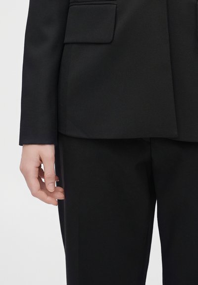 Main gauche détendue le long du corps portant un blazer noir et un pantalon noir sur un fond clair uni.