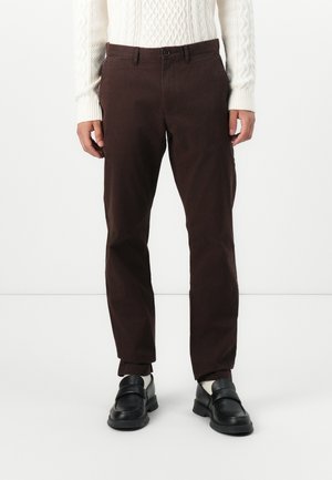 Pantaloni in cotone marroni con un design a gamba dritta, tasche laterali, e chiusura con bottone, abbinati a scarpe nere slip-on e un maglione bianco in maglia.