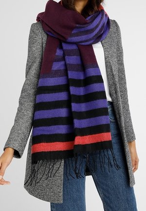 Femme portant un manteau gris, un jean bleu, un haut blanc et une écharpe épaisse à rayures horizontales noires, violettes, rouges et bordeaux.
