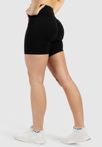 Shorts noirs taille haute à coupe ajustée, avec une texture lisse et une ceinture élastique contrastante, associés à des chaussures de sport blanches.