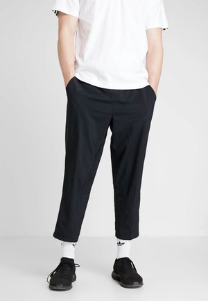 Uomo con camicia bianca a maniche corte, pantaloni neri corti, calzini bianchi Adidas e scarpe da ginnastica nere, in piedi con le mani in tasca.