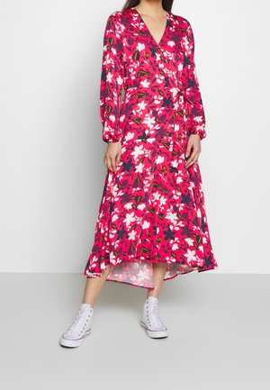 Bloemige wikkeljurk in het roze met witte en marineblauwe bloemen; heeft lange mouwen, een V-halslijn en een vloeiende zoom. Gecombineerd met witte sneakers.