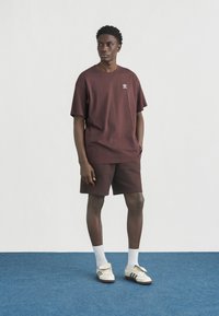 Νεαρός άνδρας που φοράει μπορντό μπλούζα και σορτς Adidas, λευκές κάλτσες και λευκά αθλητικά παπούτσια Adidas, στέκεται σε μπλε χαλί μπροστά σε λευκό τοίχο.