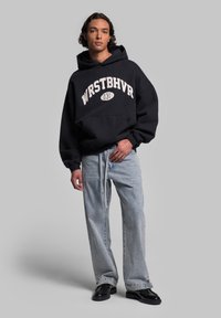 Felpa nera con logo "WRSTBHVR" in bianco, abbinata a jeans azzurri a vestibilità comoda e scarpe nere. Tessuto morbido, design casual.