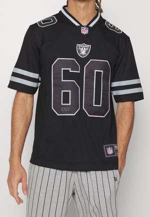 Mand iført en sort Raiders NFL fodboldtrøje med nummer 60 og grå stripede bukser, stående mod en ensfarvet baggrund.