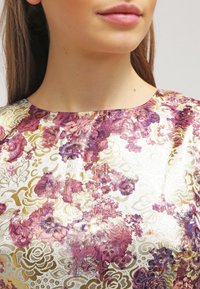 Lähikuva naisen alahuulesta ja kaulasta, joka on pukeutunut kukkakuviolliseen toppiin violettien, kullan ja valkoisten sävyjen väreissä.