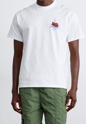 Mężczyzna w gładkiej białej koszulce z małą czerwoną grafiką wiśni i napisem na lewej piersi, w połączeniu z zielonymi spodniami bojówkami.