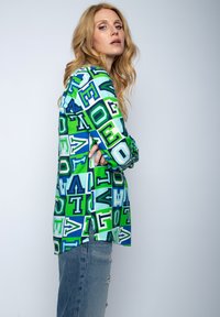 Koszula z długim rękawem z wyrazistym wzorem liter w odcieniach niebieskiego, zielonego i białego, o luźnym kroju i bocznymi rozcięciami. W połączeniu z przetartymi dżinsami.