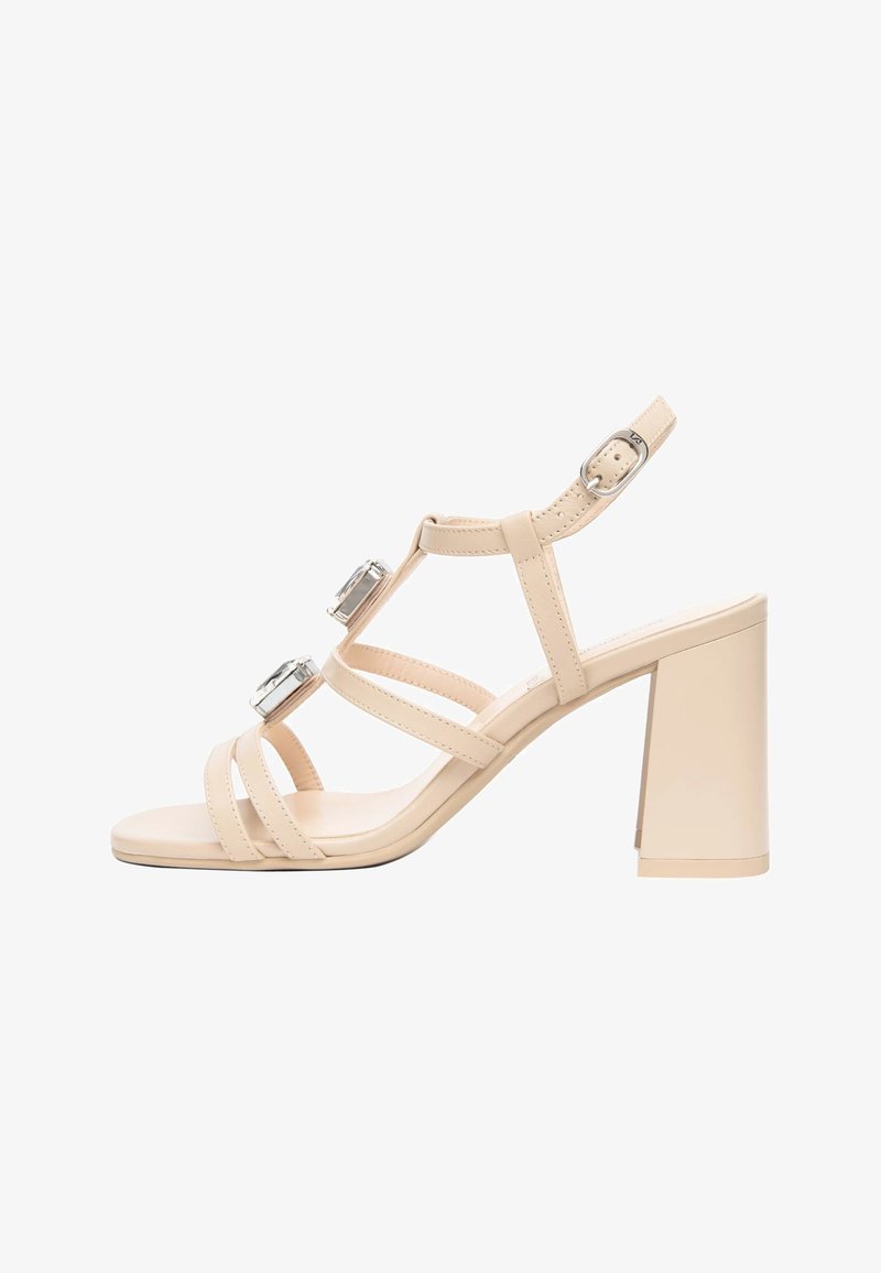 Sandale beige à talon haut avec talon bloc, trois fines brides sur le pied, deux grandes décorations carrées argentées et boucle réglable à la cheville.