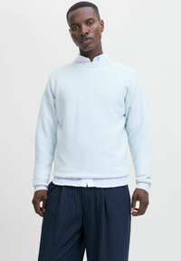 Lys blå sweater lavet af et blødt materiale med rund hals og ribbet kant. Båret over en hvid skjorte med krave og parret med stribede bukser.