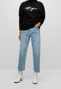 Schwarzer Sweatshirt mit weißem "Hugo"-Logo, kombiniert mit hellblauen, geraden Jeans und weißen Stiefeletten mit Blockabsatz.