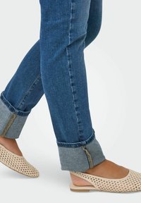 Jalat yang mengenakan celana jeans biru dengan manset dan sepatu flat slingback tenun berwarna beige di latar belakang yang terang.