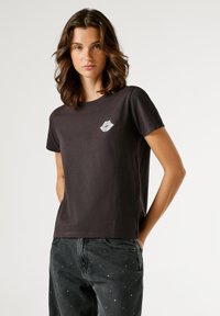 Νεαρή γυναίκα με κυματιστά καστανά μαλλιά, φορώντας ένα μαύρο t-shirt με λευκή εκτύπωση χειλιών και σκούρα τζιν με μικρές λευκές πιτσιλιές μπογιάς, με τα χέρια στις τσέπες.