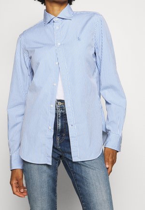 Osoba ubrana w jasnoniebieską i białą koszulę w paski zapinaną na guziki, na białym topie, oraz niebieskie dżinsy, widoczna od szyi do bioder.