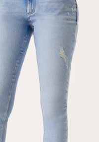Light blue distressed jeans with frayed patches and a slim fit. Features visible stitching along the seams and a smooth texture.