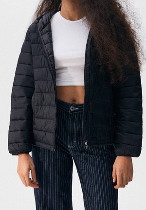 Jeune personne portant une veste matelassée noire sur un crop top blanc et un pantalon foncé à fines rayures, debout les bras détendus le long du corps.