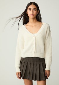 Cremfarbener Strickcardigan mit tiefem V-Ausschnitt und Knopfverschluss, kombiniert mit einem gefalteten, dunkelbraunen Rock mit dezenten Streifen.