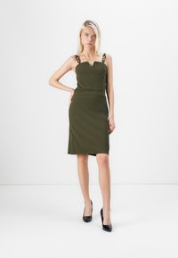 Robe vert olive avec corsage ajusté et jupe longueur genou. Présente des bretelles fines avec des accents de chaîne décoratifs et une texture lisse.