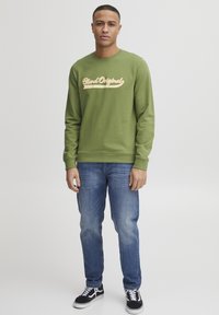 Olijfgroene sweatshirt met "Bland Original" tekst in lichtgoud, ronde halslijn, ribgebreide boorden, gecombineerd met een blauwe spijkerbroek en zwarte sneakers.