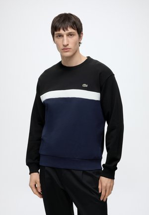 Jeune homme portant un sweat à blocs de couleurs noir, blanc et marine avec un petit logo crocodile vert sur la poitrine.