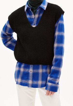 Personne portant une chemise à carreaux bleue sous un gilet sans manches en tricot noir et un pantalon blanc sur fond blanc.