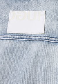 Světle modrá denimová látka s horizontálním bílým koženým štítkem s raženým nápisem. Viditelné švy podél okrajů.