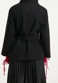 Manteau noir ceinturé à la coupe structurée, avec un col et des poignets en maille rouge. Le vêtement est assorti à une jupe noire plissée.