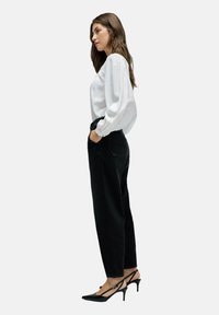 Pantalones vaqueros negros de cintura alta con un corte relajado, combinados con una blusa blanca de manga larga con puños elásticos. Unos tacones negros de punta completan el conjunto.
