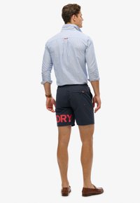 Mann trägt ein hochgekrempeltes blau gestreiftes Hemd, marineblaue Shorts mit rotem "DRY"-Schriftzug, braune Lederloafer, steht mit dem Rücken zum Betrachter vor weißem Hintergrund.