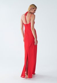 Červené večerné šaty s halter krkom, bez chrbtového dielu a bočným rozparkom. Ľahká tkanina a tenké ramienka zvyšujú jeho elegantnú siluetu.