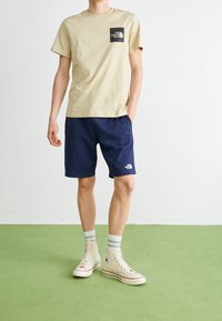 Camiseta de algodón beige con un parche de logo negro, combinada con pantalones cortos azul marino. El modelo lleva zapatillas altas beige y calcetines azul claro sobre un suelo verde.