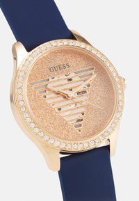 Reloj dorado con una esfera de glitter en tono rosa dorado, correa de goma azul y un bisel adornado con cristales que presenta un diseño triangular.