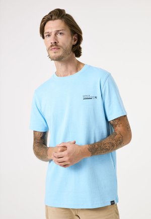 T-shirt en coton bleu clair à manches courtes, col rond, et un petit logo noir sur la poitrine gauche. Présente une coupe décontractée et une texture douce.
