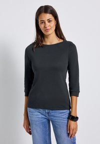 Frau mit langen, dunklen Haaren, die ein schwarzes Oberteil mit drei Viertelärmeln, hellblaue Jeans, goldene Creolen und ein schwarzes Armreifen trägt, präsentiert sich frontal.