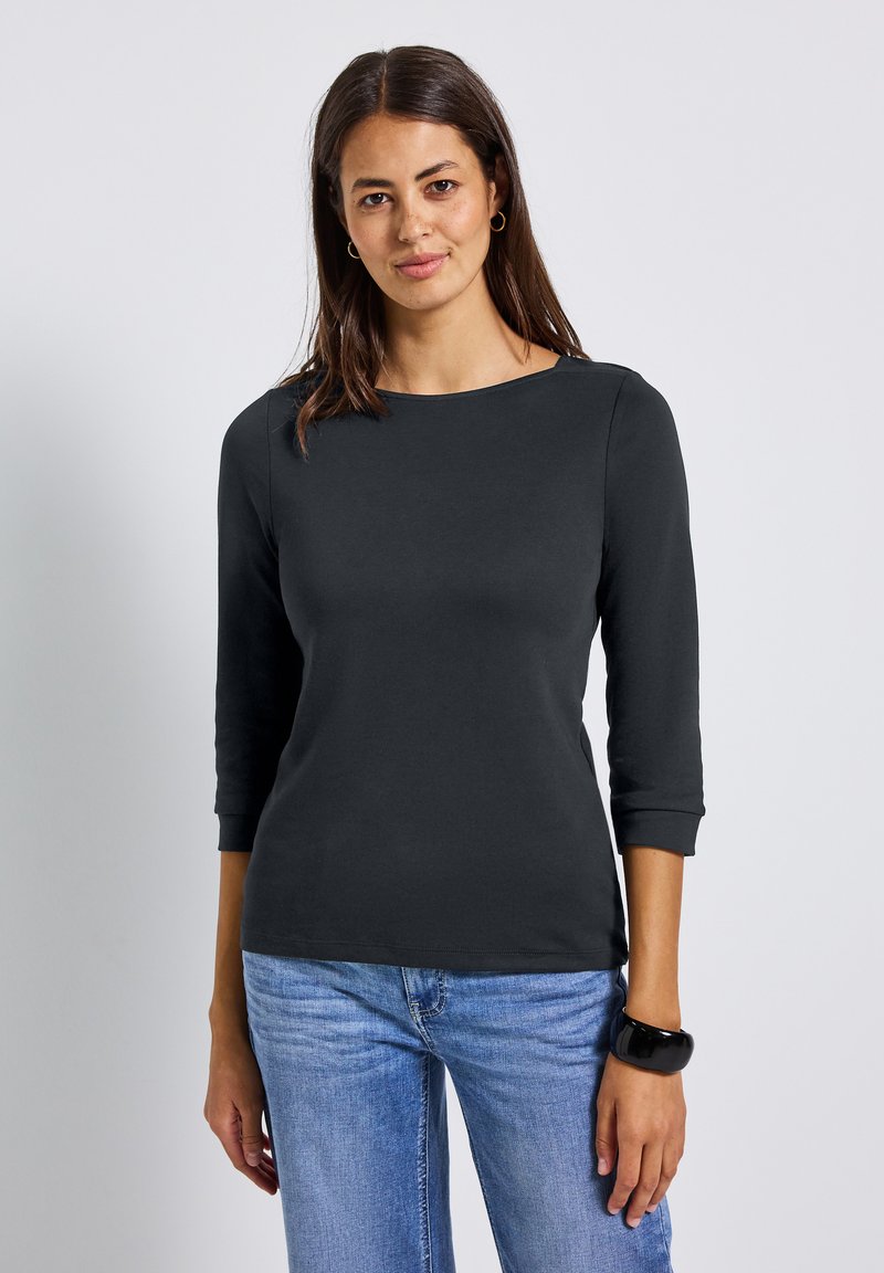 Frau mit langen, dunklen Haaren, die ein schwarzes Oberteil mit drei Viertelärmeln, hellblaue Jeans, goldene Creolen und ein schwarzes Armreifen trägt, präsentiert sich frontal.