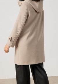 Manteau en laine beige avec une capuche, silhouette droite et poignets boutonnés. Dispose de poches latérales et d'une texture douce. Un pantalon noir complète le look.