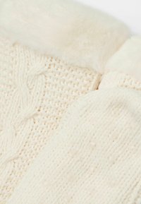 Gros plan sur un tricot de couleur crème avec des motifs en câble et en côtes, featuring des bordures douces et duveteuses au niveau des cols ou des bords.