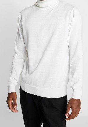 Homme portant un pull à col roulé blanc uni et un pantalon noir, debout devant un fond neutre.